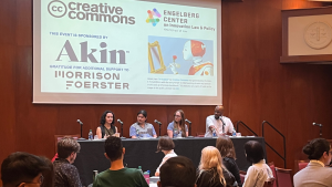People seated at table in a conference room watch a panel of four speak on stage below a slide with an image of a robot painting at an empty easel, saying: Creative Commons, Engleberg Center on Innovation Law & Policy, this event sponsored by Akin, gratitude for additional support to Morrison Foerster.